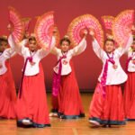 A group of dancers in traditional red and white costumes performing with pink fans on stage.