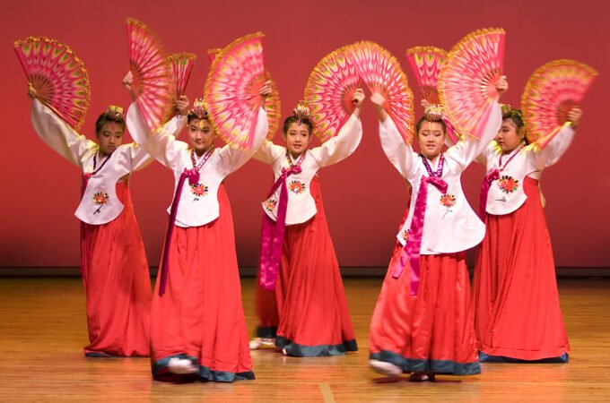 A group of dancers in traditional red and white costumes performing with pink fans on stage.