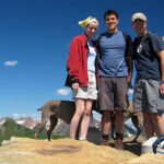 Three friends posing on a mountain top with their dog against a backdrop of snow-capped peaks and blue sky.