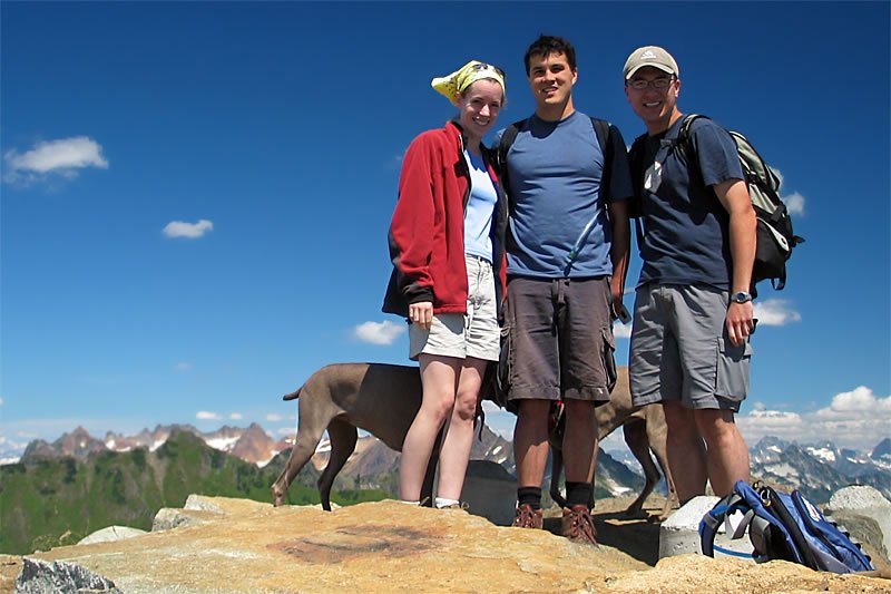 Three friends posing on a mountain top with their dog against a backdrop of snow-capped peaks and blue sky.