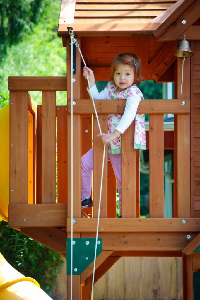 A young child climbs the rope ladder of a wooden playhouse while another person assists. The playhouse features a small bell on the roof.