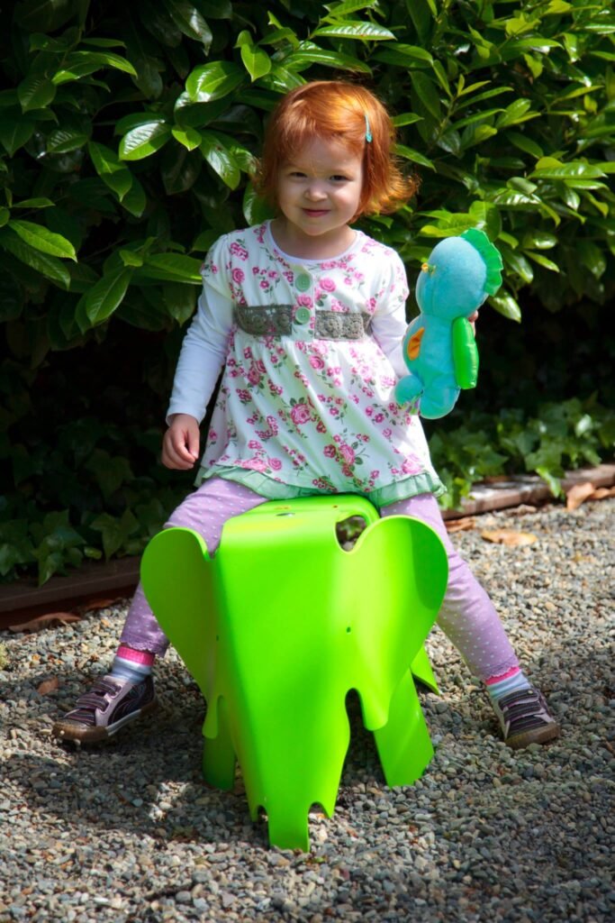 A red-haired girl sits on a bright green plastic elephant ride in a garden. She holds a green and blue balloon while wearing a floral patterned shirt.