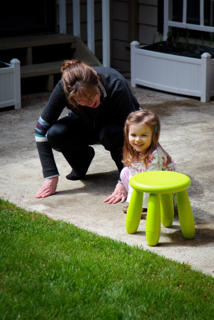 A happy child smiles next to a bright green plastic stool on a patio. An adult crouches nearby during a casual family moment on a sunny afternoon.