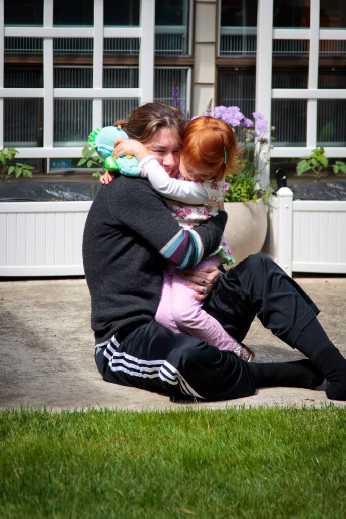 A person sits on the grass while holding and hugging two young children in front of a house. Background elements include white window frames and potted plants.