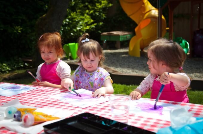 A group of children using brushes and watercolors at an outdoor table with a checkered tablecloth. Colorful paint tubes and artwork are visible on the table in a sunny, garden setting.
