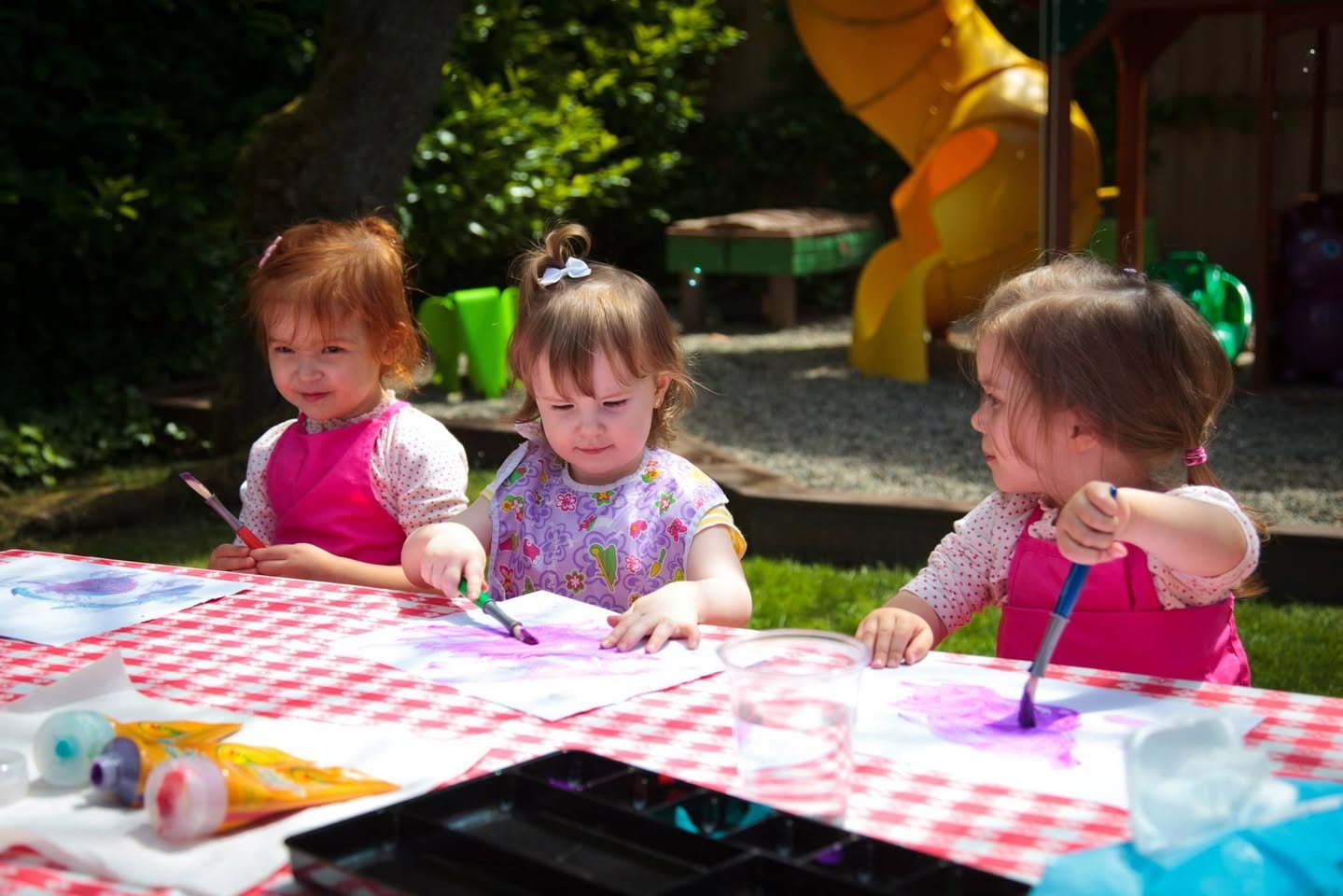 A group of children using brushes and watercolors at an outdoor table with a checkered tablecloth. Colorful paint tubes and artwork are visible on the table in a sunny, garden setting.