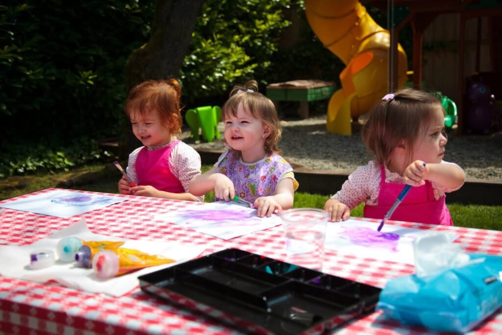 Toddler children painting with brushes on a red gingham tablecloth in a backyard. A black paint palette and squeeze bottles of paint are illuminated by bright sunlight.