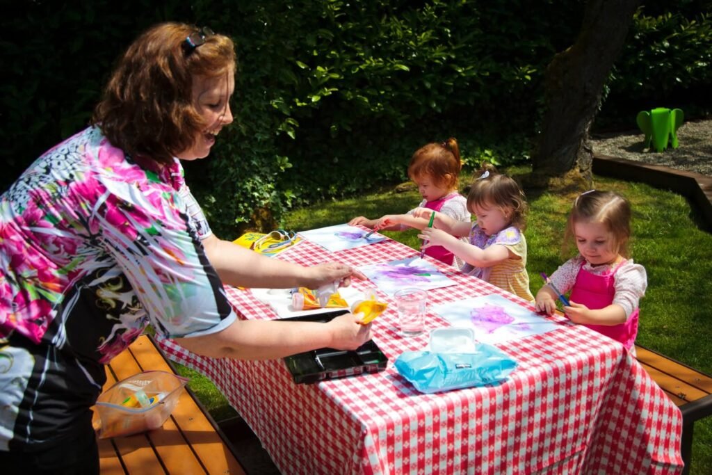 A person supervises children doing an art project at an outdoor table covered with a red and white checkered tablecloth in a grassy garden.