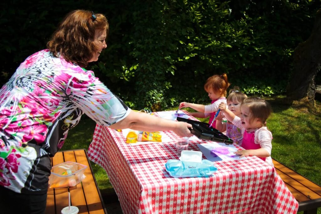 An adult and children gathered around a red checkered table with crafts in a sunny garden. Close-up of the table and seating area.