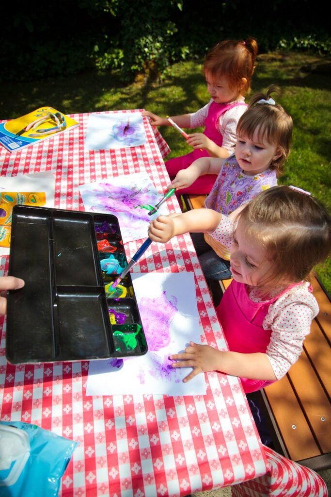 Two young children painting at a red and white checkered table outdoors. A paint palette with colorful paints is visible on the table.