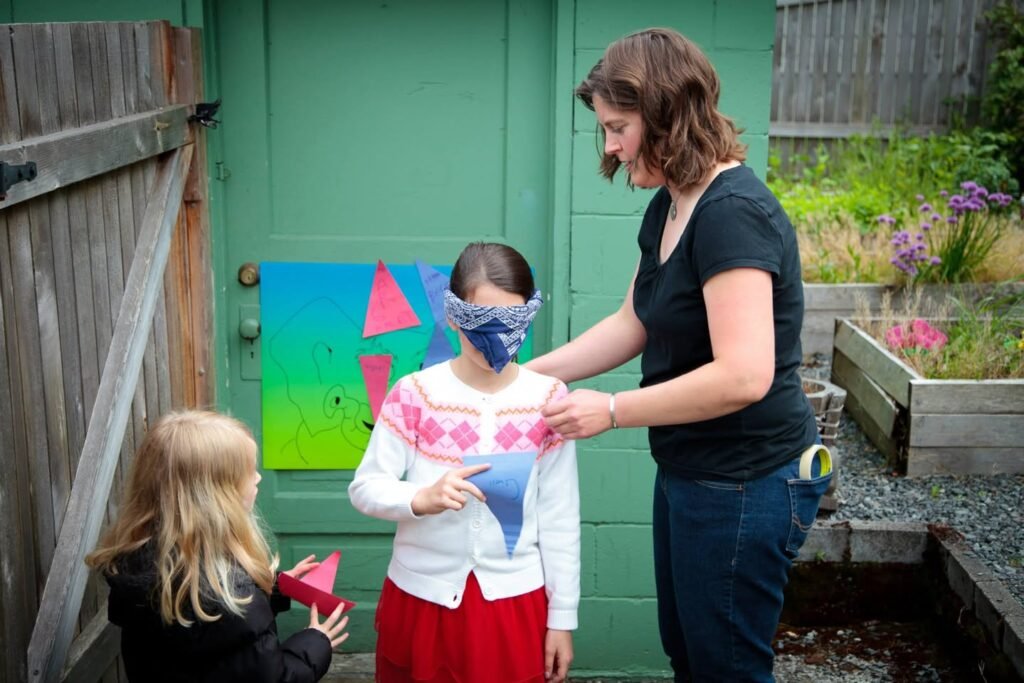 A child wearing a white knit sweater with a colorful geometric snowflake design, standing in a garden with a green wall.