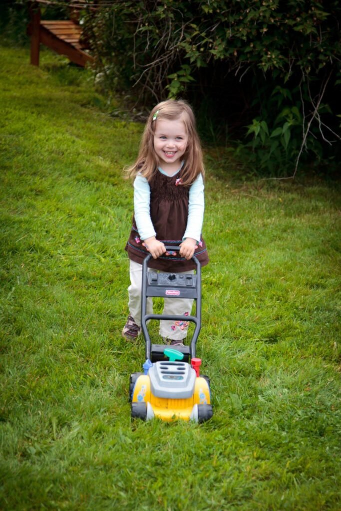 A young child pushes a yellow toy lawnmower across a green lawn. Nearby, there is a wooden bench against a background of leafy green bushes.