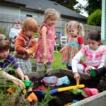 Children playing with colorful toys in a plant tray in a garden and a woman in the background.