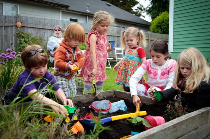 Children playing with colorful toys in a plant tray in a garden and a woman in the background.