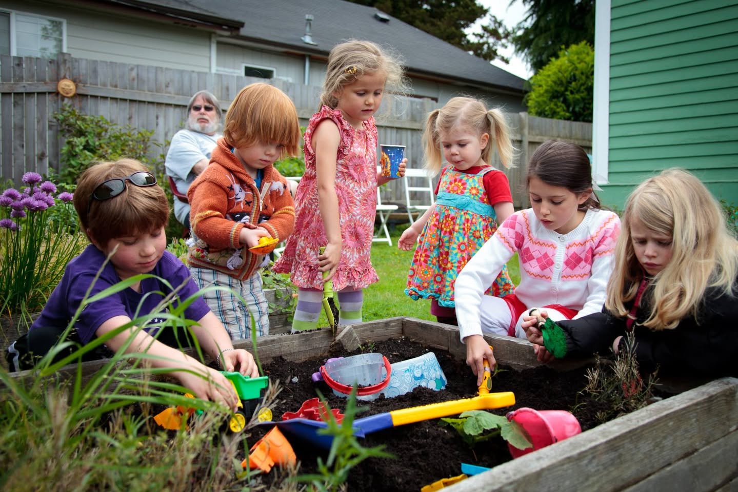 Stella & Sawyer’s 3rd Birthday Children playing with colorful toys in a plant tray in a garden and a woman in the background.