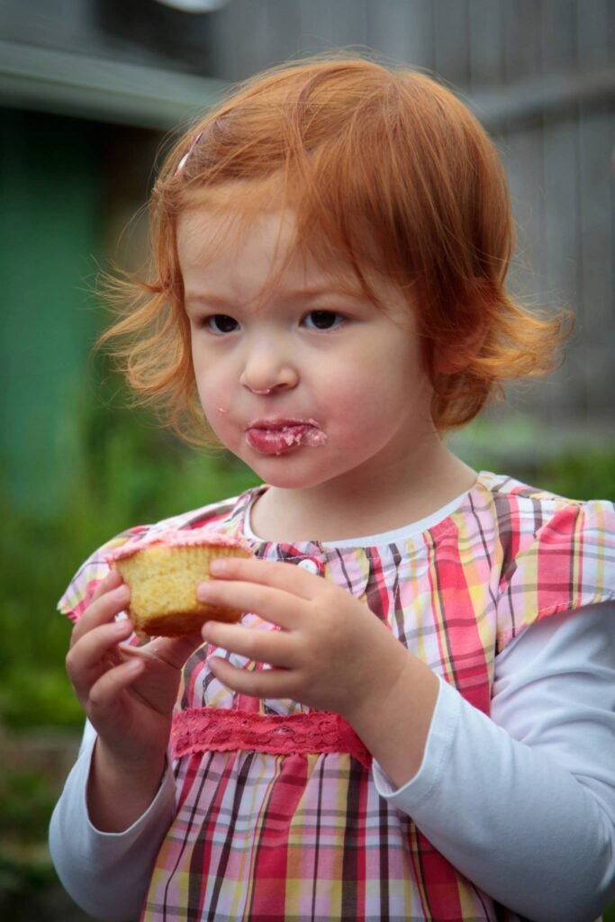 A toddler with red hair and a plaid dress holds a pancake while making a no-mouth expression while eating.