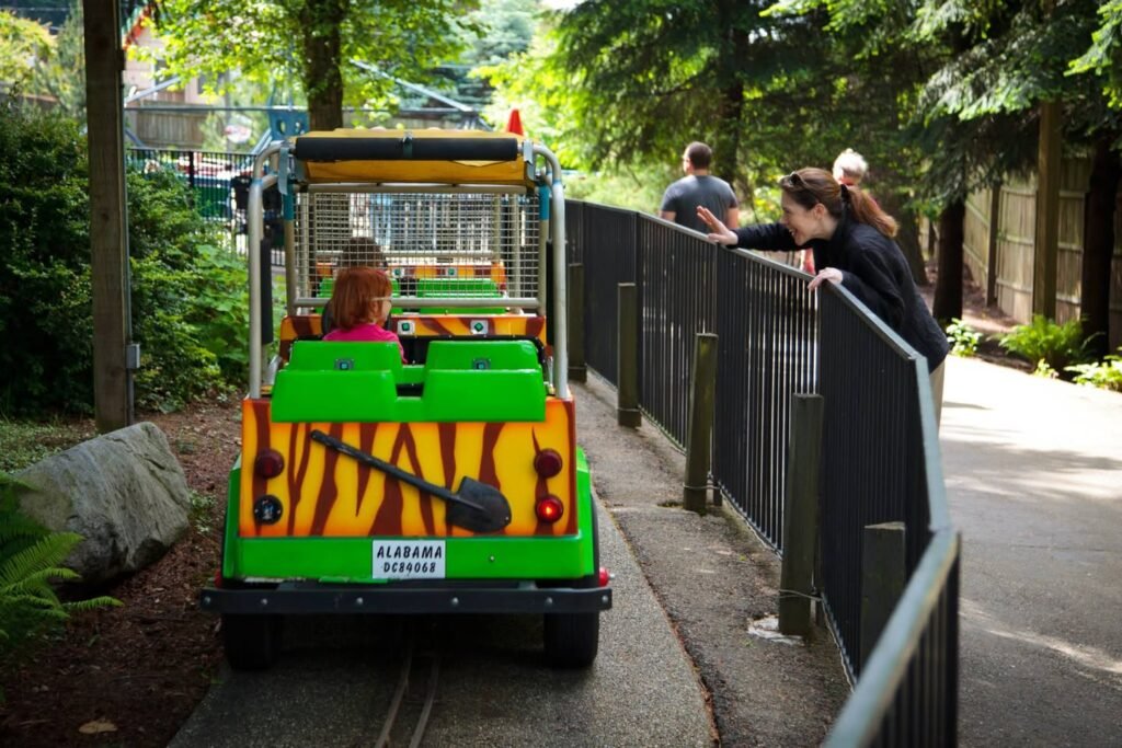 A zebra-striped mini train travels along a track in a wooded area as visitors watch from behind a railing at a zoo. Lush green foliage and natural scenery surround the scene.