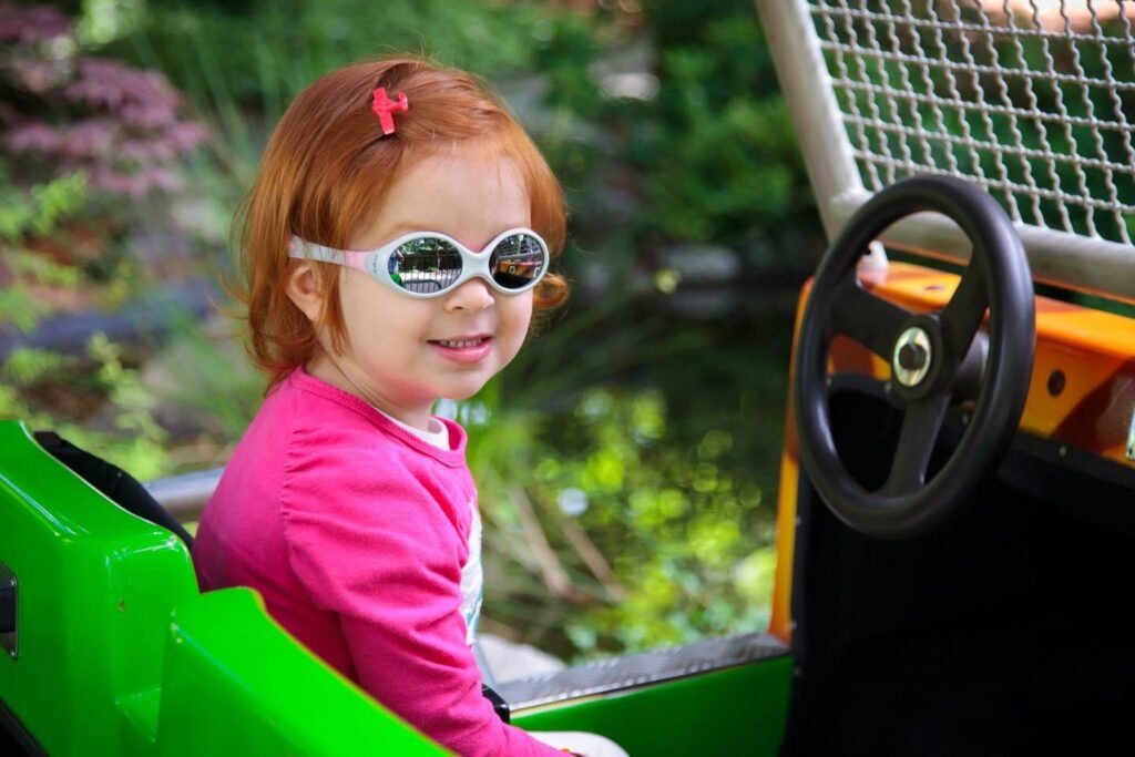 A smiling toddler in goggles and a pink shirt sits in a green toy tractor near a garden.
