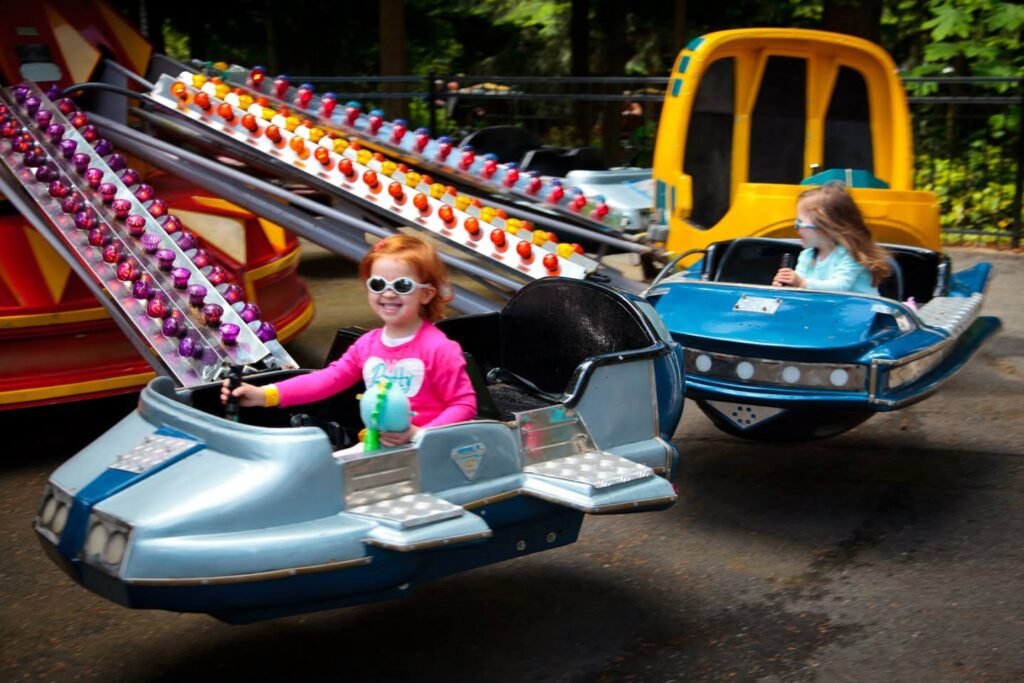 Two children are riding in simulated airplane vehicles at an amusement park, trying to hit colorful targets on a track. The scene is set outdoors with lush greenery in the background.