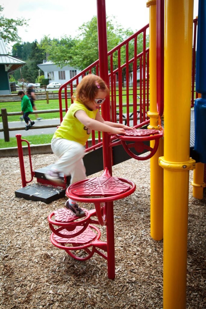 A child in a yellow top climbing red spiral stairs at a playground. A close-up shows the colorful handrails and wooden chip surface.