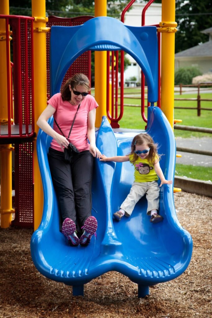 A person and a child slide down a blue playground slide together. Outdoor play equipment features bright yellow poles.