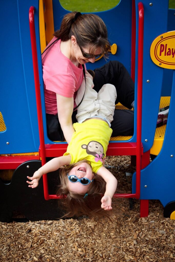 A child being lowered into wood chips by an adult at a colorful playground. The scene captures a playful moment at a 'Play' station.
