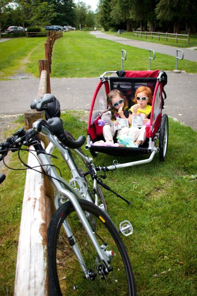 A red bike trailer with children is parked on the grass next to a bicycle in a park with a paved trail and trees in the background.