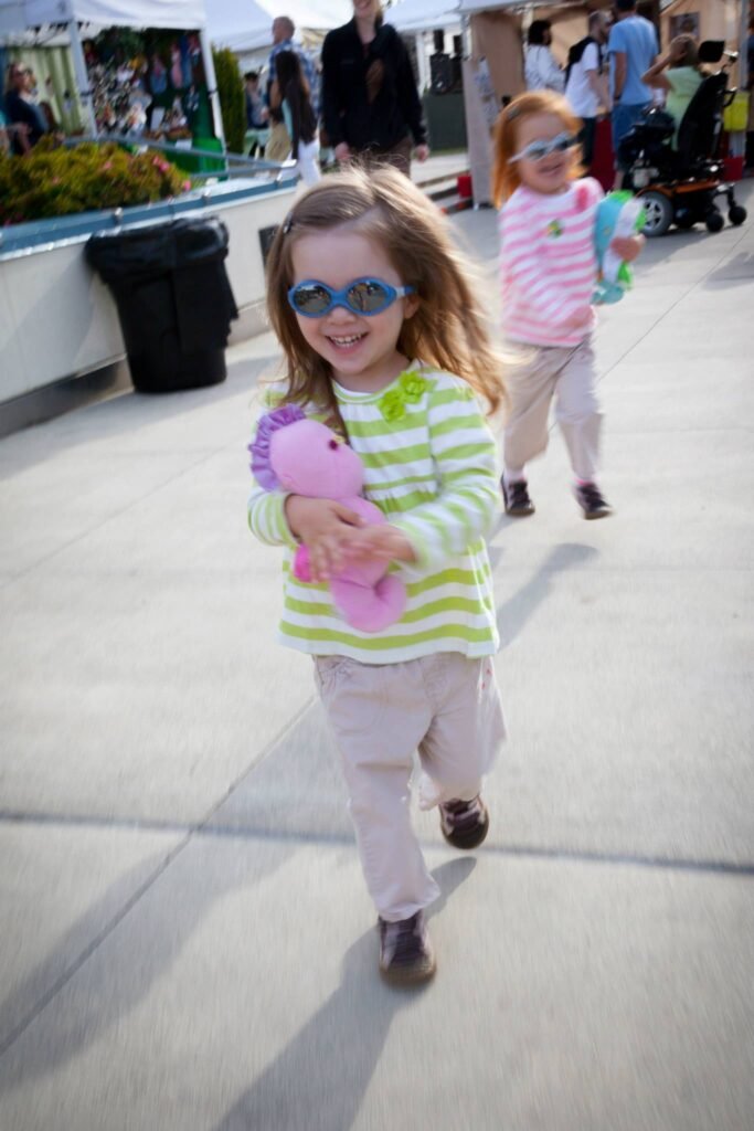 A child in a green-striped shirt holding a pink stuffed toy walks outdoors. Another younger child follows behind on a sunny sidewalk.