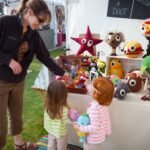 A family explores colorful handcrafted plush toys at an outdoor market.