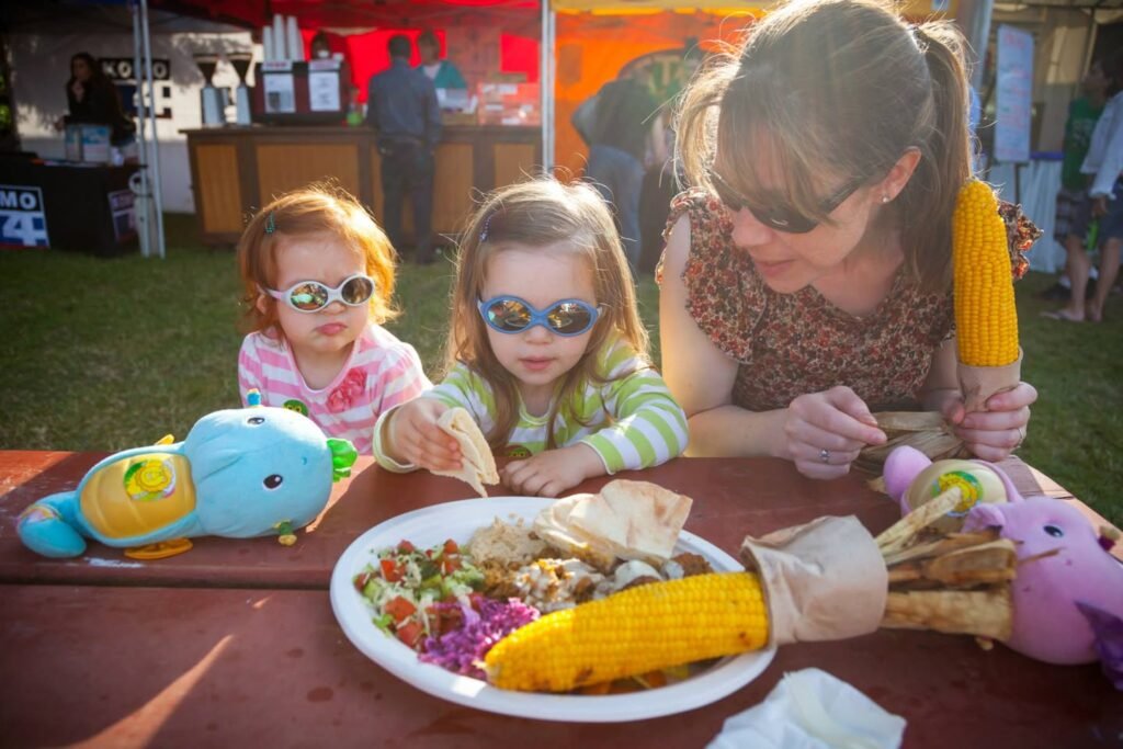 A family sits around a picnic table at an outdoor event sharing a meal of corn on the cob, rice, and salad.