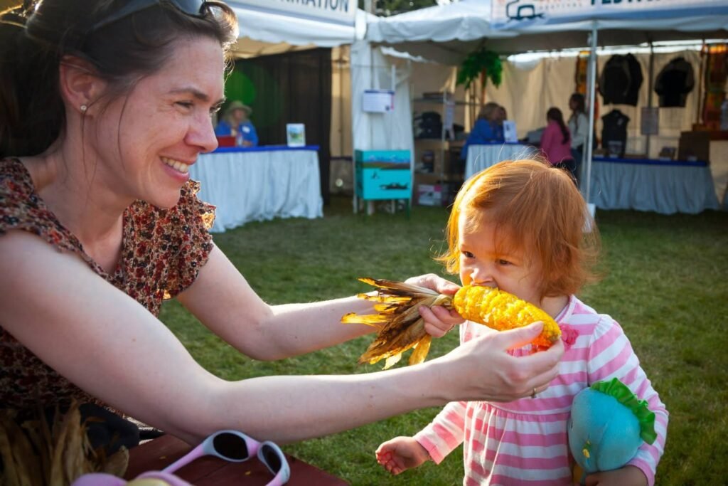 A woman offering an ear of corn to a young child at an outdoor farm event.