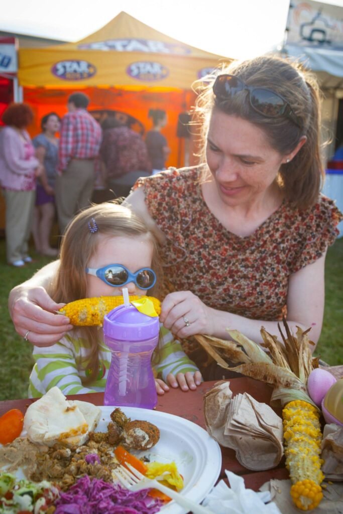 A smiling adult and a young child with sunglasses enjoying fresh corn at an outdoor farmers market.