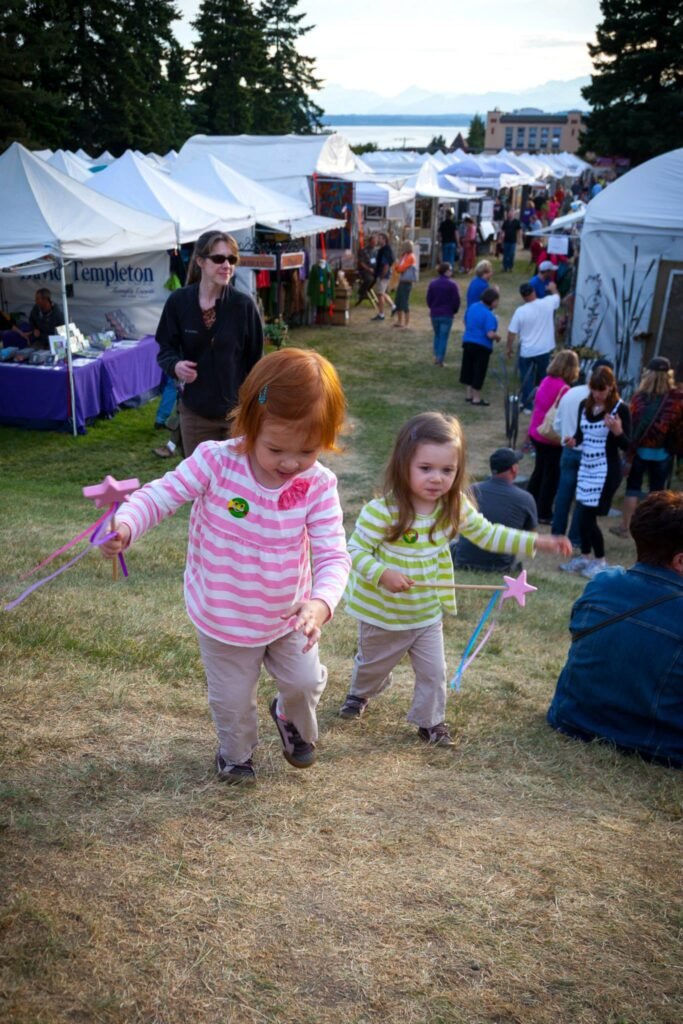 Two children play with a pinwheel at an outdoor town fair featuring market tents.