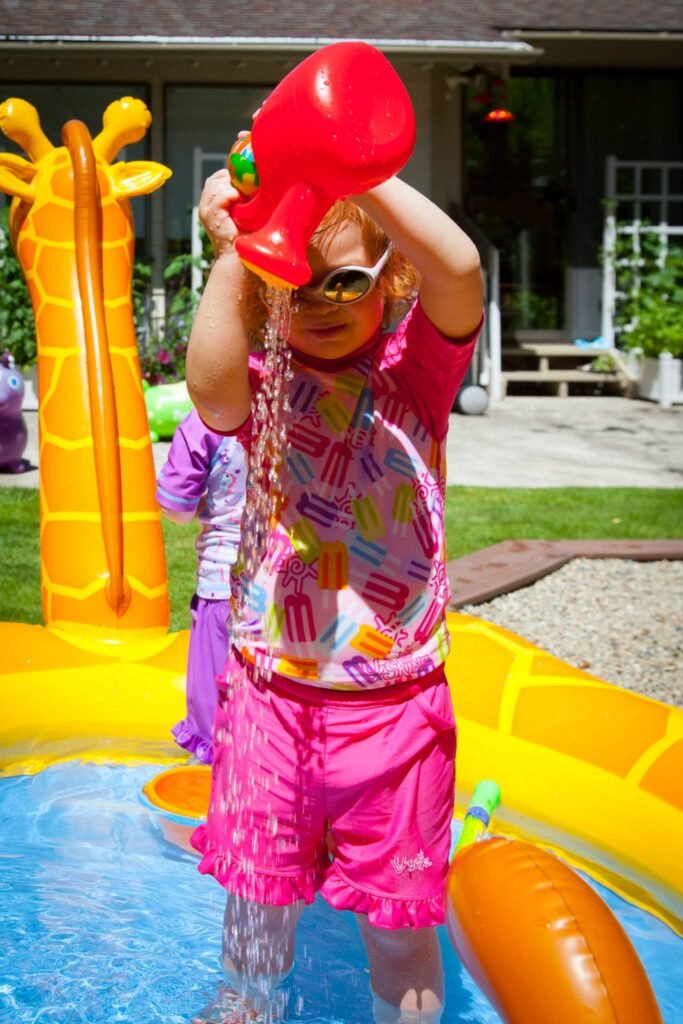 A child playing in an inflatable pool with a red water toy. The scene captures a sunny day of outdoor water play.