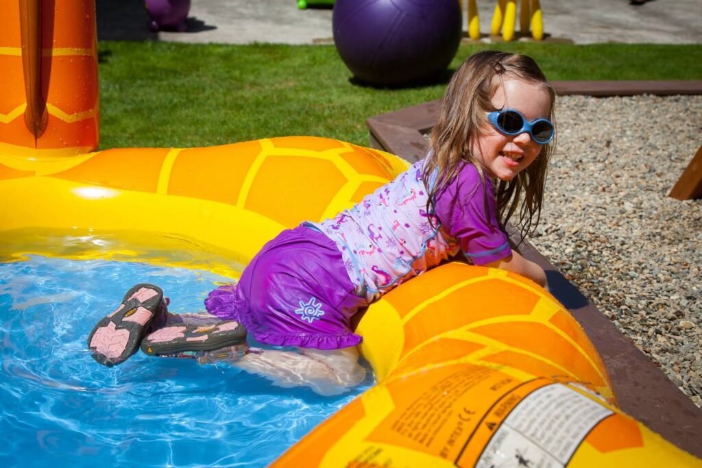 A child in purple swimsuit and sunglasses smiles in a yellow inflatable kiddie pool.