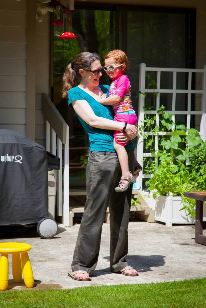 A person holding a small child in pink on a sunny backyard patio with green plants in the background.