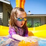 Toddler in colorful goggles plays in a bright yellow bubble pool outdoors.