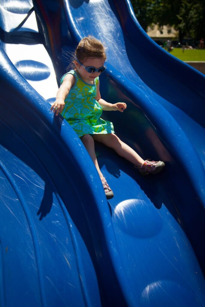 A toddler in a green dress and sunglasses slides down a large blue plastic slide on a sunny day.