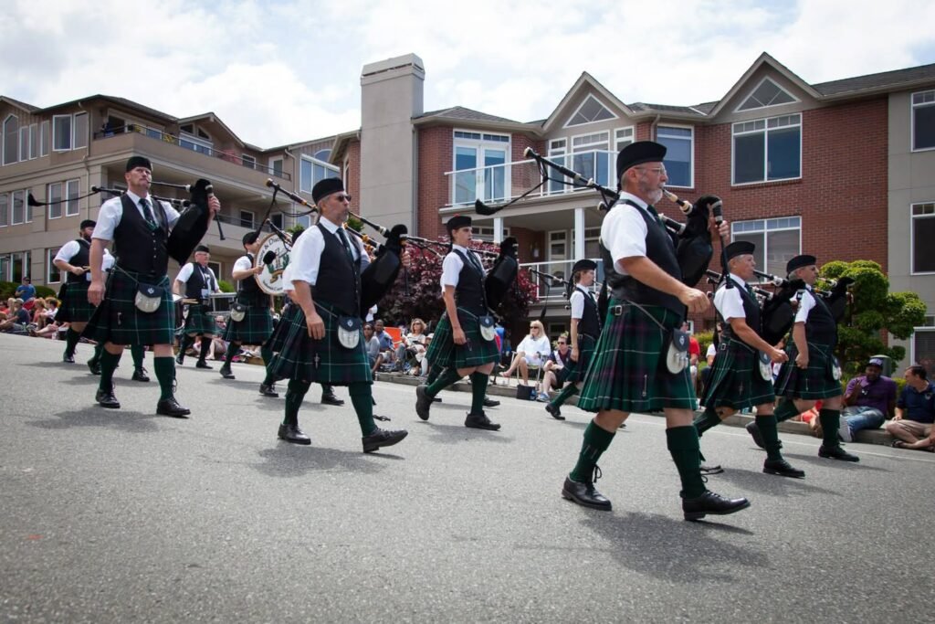 A group of bagpipers dressed in traditional kilts march down a residential street lined with spectators and townhome-style buildings.