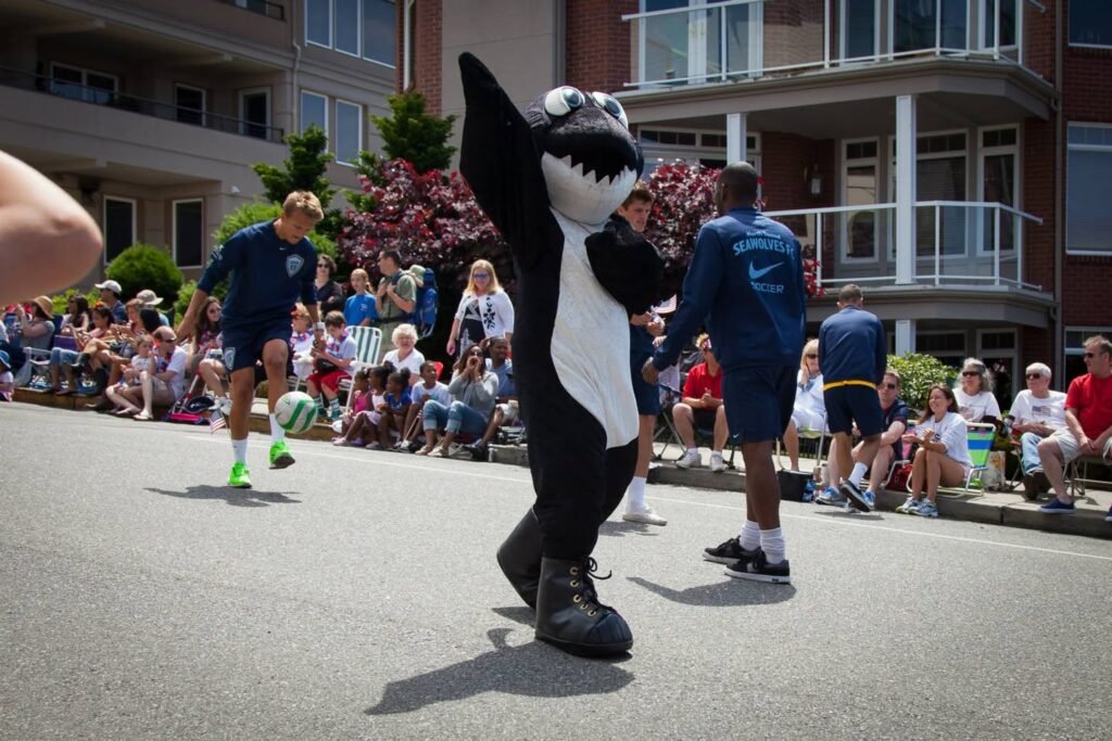 A person dressed as a penguin with a shark head dances on a city street during a soccer match in front of a cheering crowd.