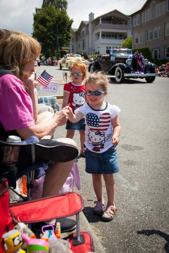 Children in patriotic shirts watch a classic car parade on a sunny day in the Pacific Northwest.