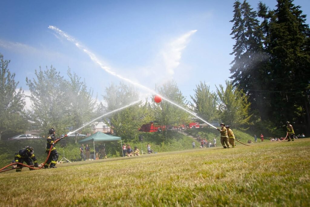 Firefighters spraying water hoses at a community event. Water arcs through the air against a backdrop of evergreen trees and a clear blue sky.