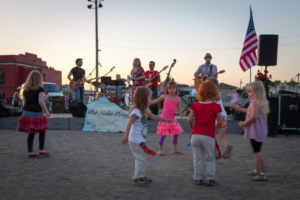 A group of young children dance in a circle at an outdoor community event with a local band playing in the background.