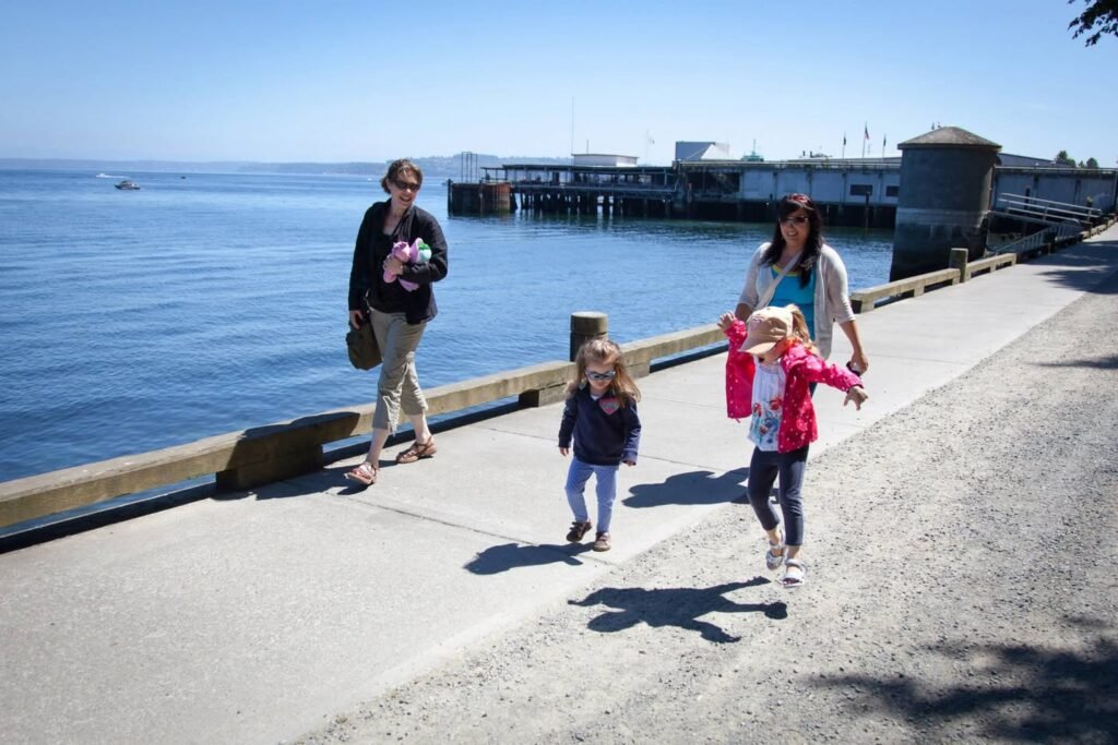 A family enjoys a sunny day at the waterfront, with children skating and walking near a historic pier.