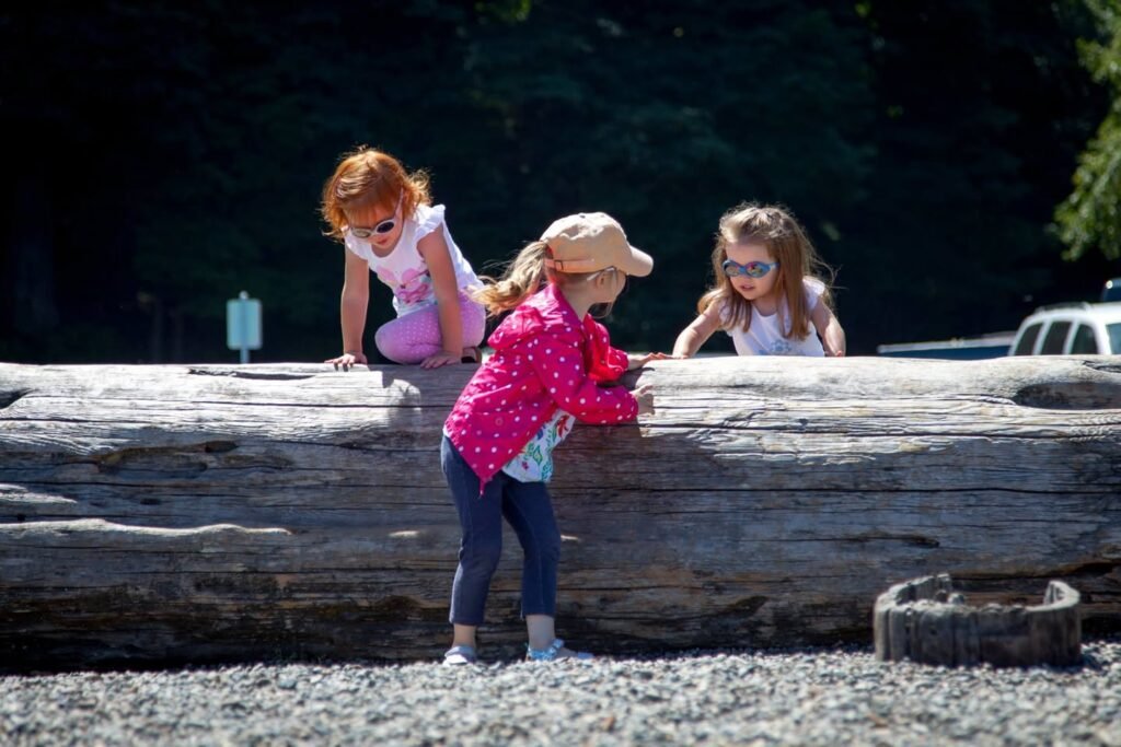Three young girls play on a large fallen tree trunk at a gravel beach. One wears a bright pink shirt and hat, exploring nature.