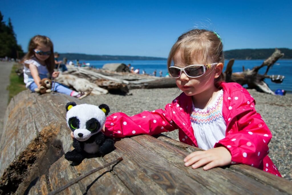 A child in a pink coat relaxes on a log at a beach with a small black and white plush panda beside them. The background features blue water, driftwood, and a clear sky.