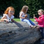 Children playing with toys on a large log in a sunny park.