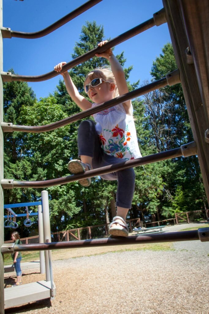 A child climbs a metal play structure at a park with tall green trees in the background.