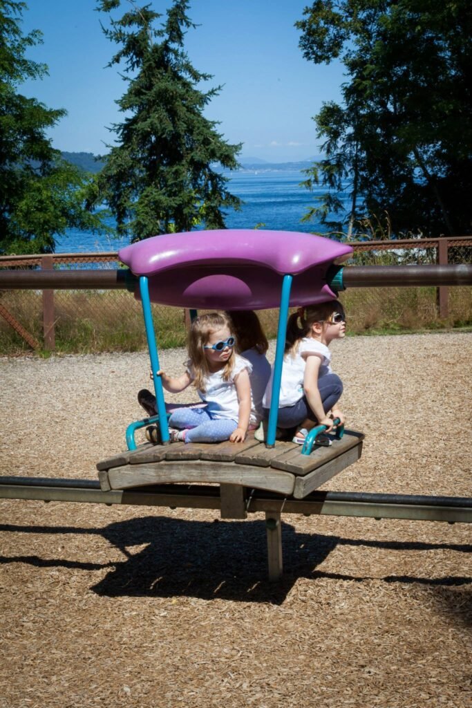 Three children enjoying a purple amusement ride in a gravel park near a large body of water, with tall green trees in the background.