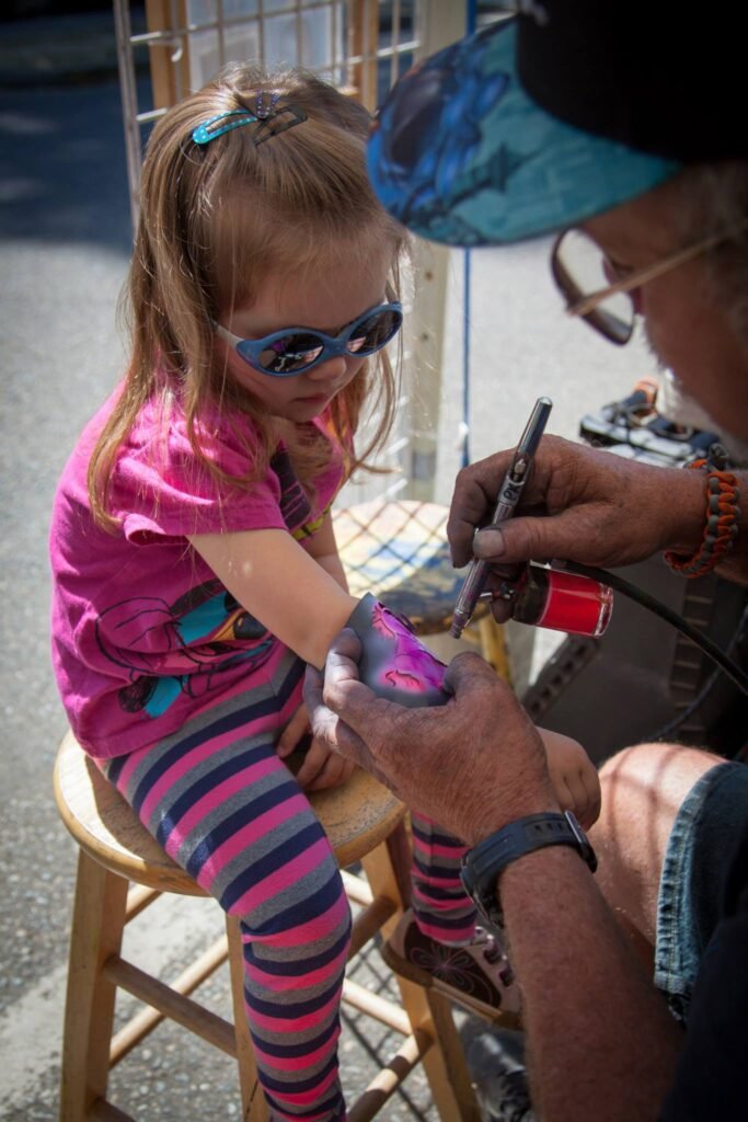 A young visitor receives a small tattoo on her arm while wearing bright sunglasses and striped leggings. An artist carefully applies the ink during a casual outdoor session.